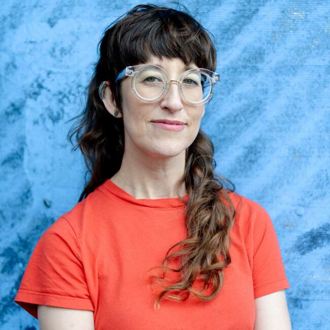Headshot of filmmaker Alison O'Daniel, in glasses, chest length hair and tangerine colored t shirt against blue background.