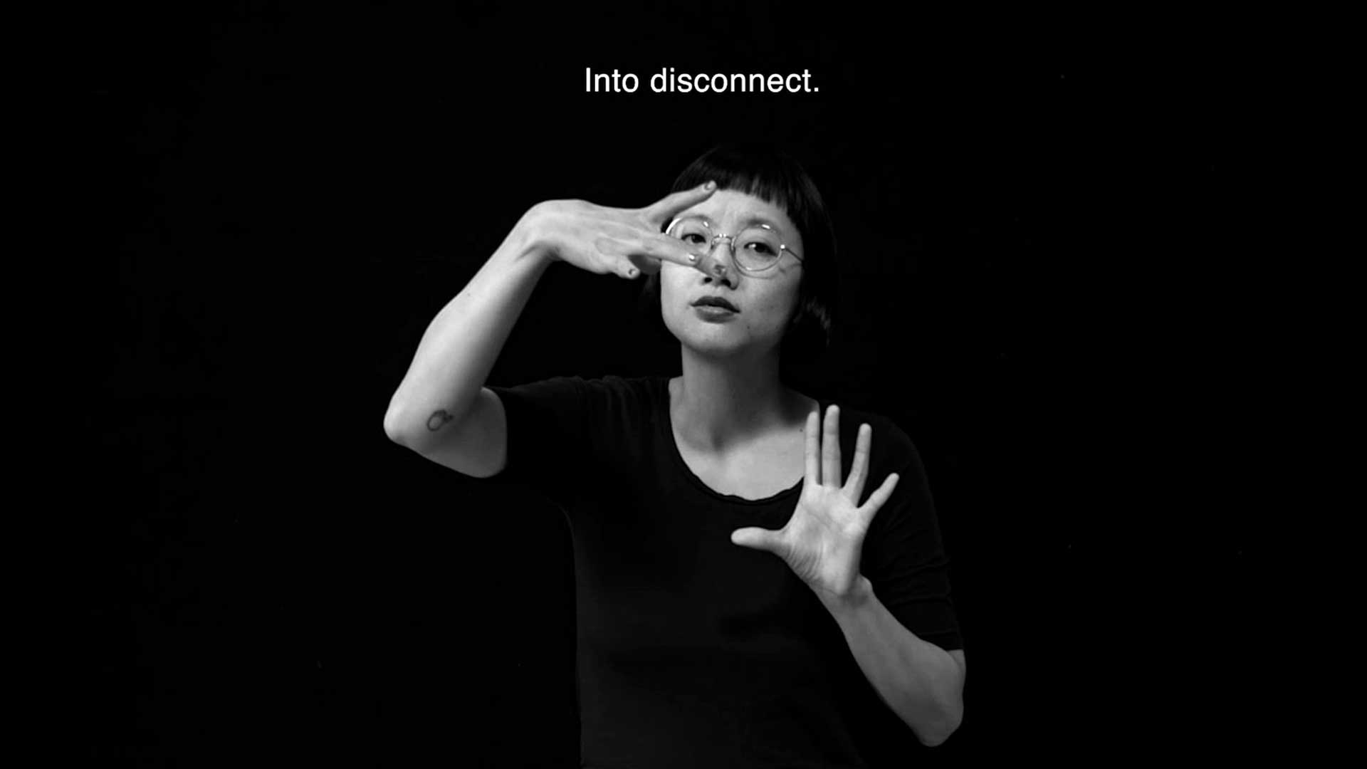 Christine Sun Kim, a Korean American womanwith black bangs, round glasses and wearing black clothing that blends into a black background, signs in American Sign Language the word 'disconnect.’ Above her a white caption says "Into disconnect." Alt text: Black and white image of a woman signing in American Sign Language the word ‘disconnect.’ The caption reads “Into disconnect.”